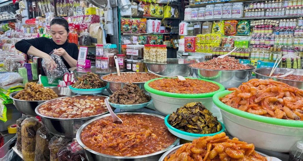 Hình ảnh gian hàng đặc sản tại chợ truyền thống Việt Nam bày bán nhiều loại mắm, tôm khô, cá khô, mực khô, hải sản chế biến và gia vị đặc trưng. Đây là điểm đến lý tưởng để du khách khám phá văn hóa ẩm thực địa phương và mua đặc sản làm quà.